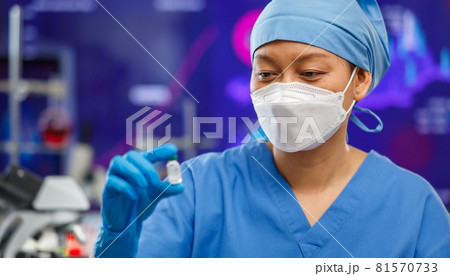 Female medical people wearing a hygiene face mask and covered clothes staying in laboratory room, holding and looking to a bottle of Covid-19 vaccine with lab tools in background. Focus on eyes Female medical people wearing a hygiene face mask and covered clothes staying in laboratory room, holding and looking to a bottle of Covid-19 vaccine with lab tools in background. Focus on eyes 81570733