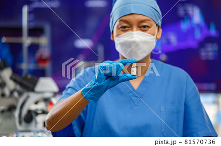 Female medical people wearing a hygiene face mask and covered clothes staying in laboratory room and holding bottle of Covid-19 vaccine with lab tools in background. Selective focus. 81570736