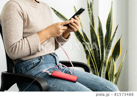 The hand of a young woman holding a smart phone and a modern spare battery charger Place the lap to charge the smartphone with a spare battery charger and play on a chair in room 81573488