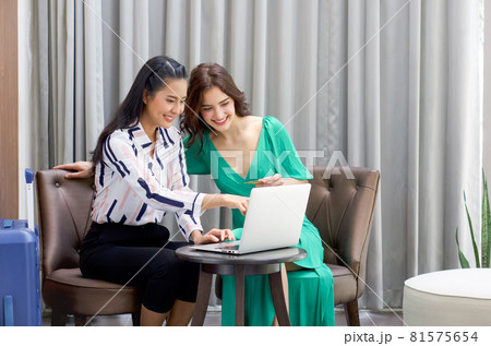 Young woman with luggage pointing at laptop computer screen, show her friend the product they looking for on internet. Caucasian woman holding credit card sitting beside her friend in hotel lobby. 81575654