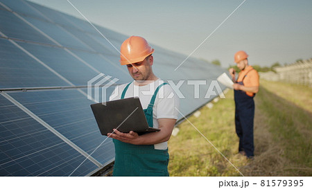 Engineer standing with Solar cell panels looking monitor of the Electric power value made from the solar cell. Man holding laptop in her hands standing on field with solar panels. 81579395