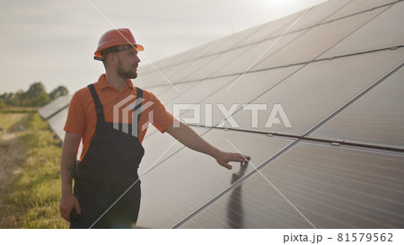 Portrait of happy male engineer in protective helmet, He speaks on a walkie-talkie while looking to camera. Handsome man in uniform smiling while standing at solar power farm. Concept of green energy Portrait of happy male engineer in protective helmet, He speaks on a walkie-talkie while looking to camera. Handsome man in uniform smiling while standing at solar power farm. Concept of green energy 81579562