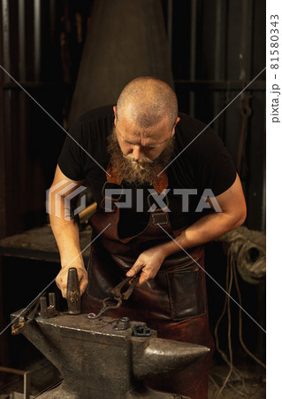 Bearded man, blacksmith manually forging the molten metal on the anvil in smithy with spark fireworks. Concept of labor, retro professions, family business 81580343