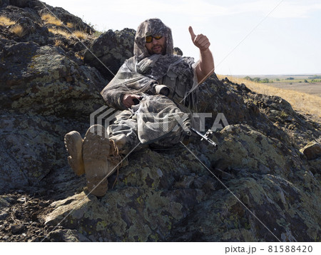 Photo of a hired sniper in camouflage during rest. He sits on the rocks and shows his thumbs up. Photo of a hired sniper in camouflage during rest. He sits on the rocks and shows his thumbs up. 81588420