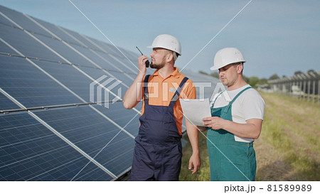 Coworking engineers on solar plant. Adult men in hardhats, An employee of a power plant transmits commands by walkie-talkie. Transportation of clean energy. Clean energy. Sunny batteries. 81589989