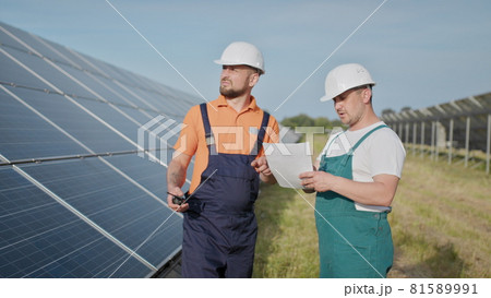 Coworking engineers on solar plant. Adult men in hardhats, An employee of a power plant transmits commands by walkie-talkie. Transportation of clean energy. Clean energy. Sunny batteries. 81589991