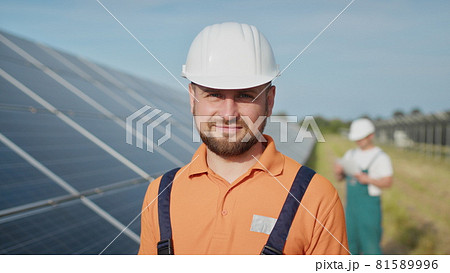 Happy caucasian adult engineer in helmet smiling at camera at solar power station outside. Ecological industry. Solar farm concept. An employee of a power plant transmits commands by walkie-talkie Happy caucasian adult engineer in helmet smiling at camera at solar power station outside. Ecological industry. Solar farm concept. An employee of a power plant transmits commands by walkie-talkie 81589996