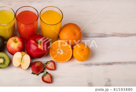 Different fruits juice in glass, apple, orange and strawberry juice with straw, looking refreshing on colourful wood board in front of white wall background. Picture decorating with fresh fruits Different fruits juice in glass, apple, orange and strawberry juice with straw, looking refreshing on colourful wood board in front of white wall background. Picture decorating with fresh fruits 81593880