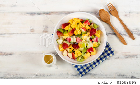 Mixed fruits salad including strawberry, kiwi, apple, and pineapple in white dish place on sackcloth on wooded table. Utensil spoon and fork and oil salad dressing cup beside Mixed fruits salad including strawberry, kiwi, apple, and pineapple in white dish place on sackcloth on wooded table. Utensil spoon and fork and oil salad dressing cup beside 81593998