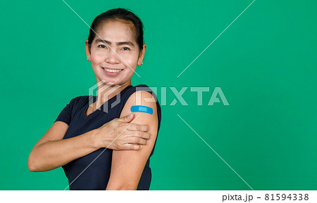 Middle aged Asian woman smiling showing her arm with bandage patch showing she got vaccinated for Covid 19 virus on green background. Concept for Covid 19 vaccination Middle aged Asian woman smiling showing her arm with bandage patch showing she got vaccinated for Covid 19 virus on green background. Concept for Covid 19 vaccination 81594338