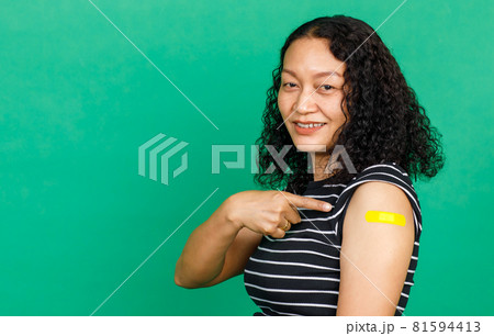 Asian middle aged woman smiling and pointing at her arm with bandage patch showing she got vaccinated for Covid 19 virus on green background. Concept for Covid 19 vaccination 81594413