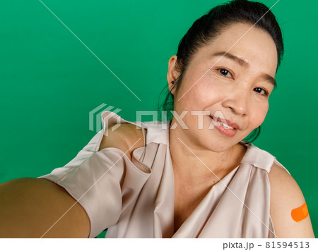 Asian middle aged woman taking selfie showing her arm with bandage patch that got vaccinated for Covid 19 virus on green background. Concept for Covid 19 vaccination 81594513