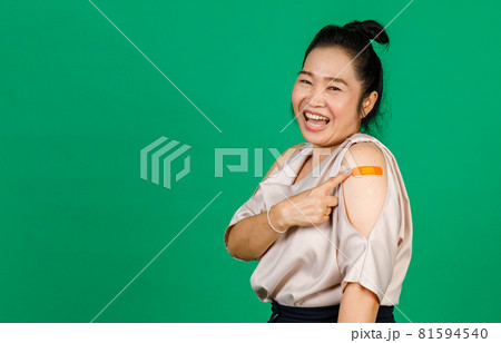 Asian middle aged woman smiling and pointing at her arm with bandage patch showing she got vaccinated for Covid 19 virus on green background. Concept for Covid 19 vaccination 81594540