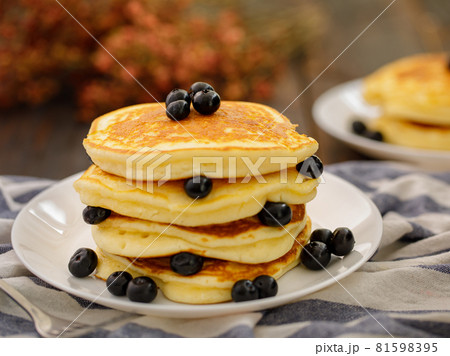 Stack of delicious pancakes sweet food with blueberries and blur dry leaves, plaid fabric in white plate on wooden background. 81598395