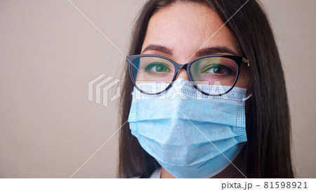 Close up of young brunette woman doctor with glasses and light blue protective mask Close up of young brunette woman doctor with glasses and light blue protective mask 81598921