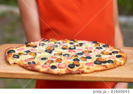 A girl in an orange t-shirt is holding homemade pizza on wooden board outdoors. Slice of pizza with shrimp, mussels, black olives and mozzarella. Home Holiday Cooking. Closeup, selective focus 81609043