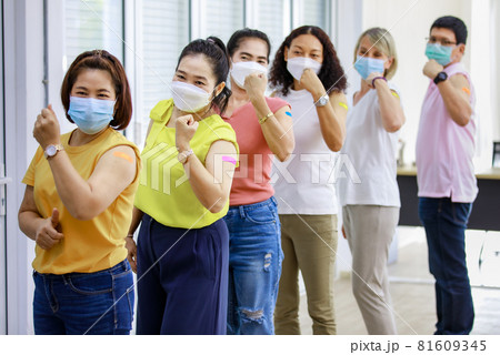 Close up shot of Asian happy strong female patient wears face mask stand look at camera in front of vaccinated orderly queue line in blurred background showing thumb up and bicep muscle up together 81609345