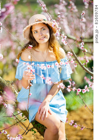 Portrait of an attractive girl in blue dress and hat in blooming peach garden Portrait of an attractive girl in blue dress and hat in blooming peach garden 81609784