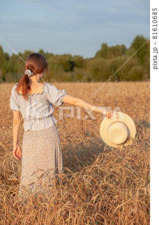 Young beautiful girl with long curly hair poses in a wheat field in the summer at sunset. A girl holds a hat in her hand against the background of a wheat field. The view from the back. Toning. 81610685