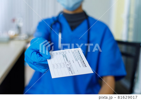 Closeup shot of Covid-19 vaccination record card certificate passport paper sent to patient by doctor wears blue hospital uniform face mask rubber gloves and stethoscope in blurred background Closeup shot of Covid-19 vaccination record card certificate passport paper sent to patient by doctor wears blue hospital uniform face mask rubber gloves and stethoscope in blurred background 81629017