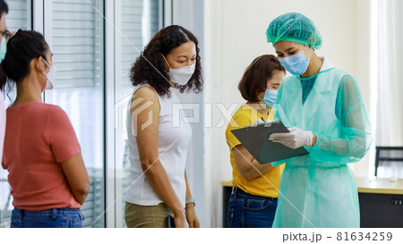Young female nurse in full hazard protection uniform holding paper board writing and asking personal information from senior curly hair woman patient in coronavirus vaccinating queue line 81634259