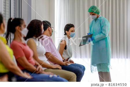 Young female citizen wears face mask in Coronavirus vaccinating queue line sitting checking her personal information from paper board in hand of full protective suit nurse who standing nearby Young female citizen wears face mask in Coronavirus vaccinating queue line sitting checking her personal information from paper board in hand of full protective suit nurse who standing nearby 81634550