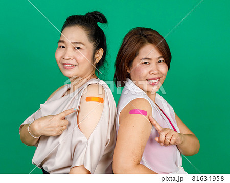 Two middle aged Asian women showing their arms with bandage patch showing they got vaccinated for Covid 19 virus on green background. Concept for Covid 19 vaccination Two middle aged Asian women showing their arms with bandage patch showing they got vaccinated for Covid 19 virus on green background. Concept for Covid 19 vaccination 81634958