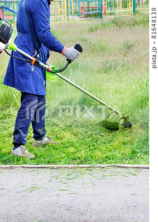 A worker in a blue overalls mows green grass with a gasoline trimmer. Vertical image. A worker in a blue overalls mows green grass with a gasoline trimmer. Vertical image. 81648139