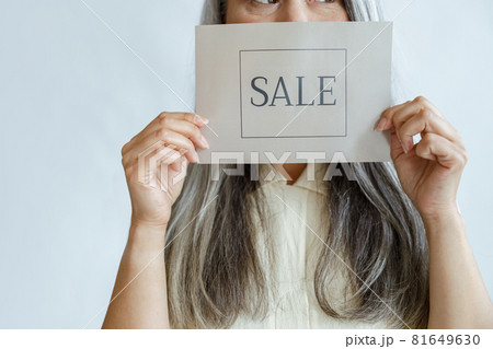 Middle aged lady with loose grey hair holds Sale sign near face standing in studio 81649630