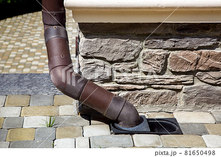 downpipe made of brown pvc plastic drainage of rainwater from roof of house into ground drainage system on corner of facade of building lined with natural stone with sidewalk made of tiles. 81650498