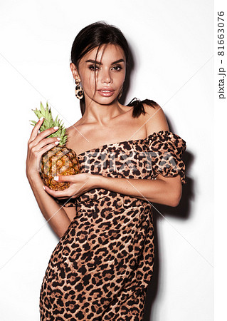 Beautiful sexy woman with pineapple in hands in a leopard dress and earrings, with classic smokey makeup and a fashionable hairstyle. Beauty face. 81663076