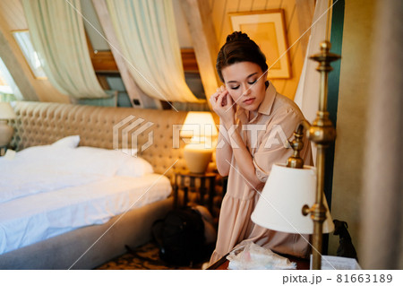 a beautiful young woman puts on earrings by the mirror in the bedroom.  81663189