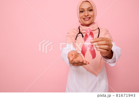 Blurred Muslim doctor in pink hijab posing on pink background, showing support, solidarity to patients cancer survivors. World Day of fight cancer. Focus on Pink Breast Cancer Awareness ribbon 81671943