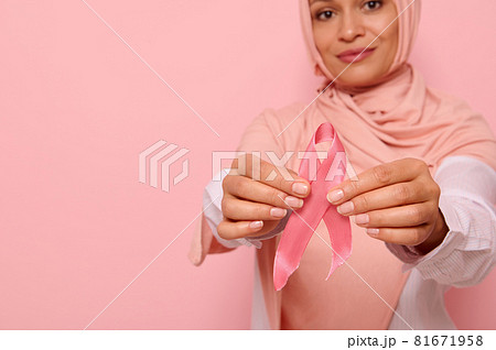 Focus on pink Breast Cancer Awareness ribbon in the manicured hands of blurred beautiful friendly Arab Muslim woman in pink hijab headscarf on pink background with copy space. World Cancer Day concept 81671958