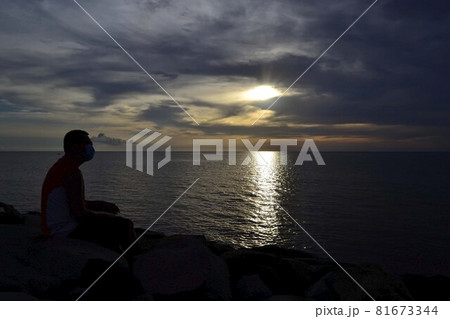 Man wearing facemask sitting on a rock watching the sunset at sea Man wearing facemask sitting on a rock watching the sunset at sea 81673344
