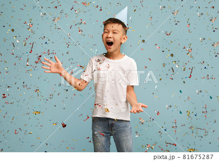 Birthday Party. Happy Asian Boy Wearing B-Day Hat Standing Under Falling Confetti Birthday Party. Happy Asian Boy Wearing B-Day Hat Standing Under Falling Confetti 81675287