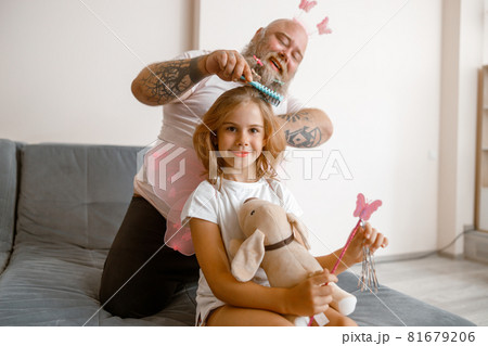 Smiling girl holds toy dog and magic stick while dad brushes her hair in room Smiling girl holds toy dog and magic stick while dad brushes her hair in room 81679206