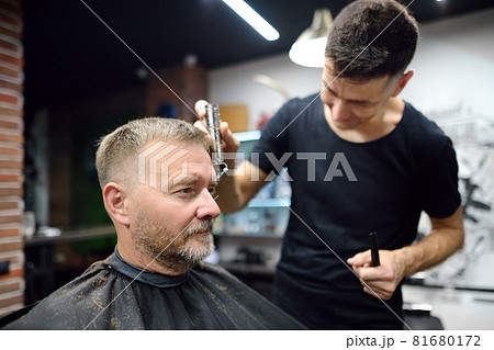 Hairdresser is doing eyebrow trimming using a razor for handsome bearded mature man in salon. Stylist making hairstyle for client in barbershop. Services of a professional stylist. Hairdresser is doing eyebrow trimming using a razor for handsome bearded mature man in salon. Stylist making hairstyle for client in barbershop. Services of a professional stylist. 81680172