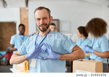 Portrait of caucasian male volunteer in blue uniform and protective gloves showing heart love sign, looking at camera. Team sorting, packing items in cardboard boxes 81681656