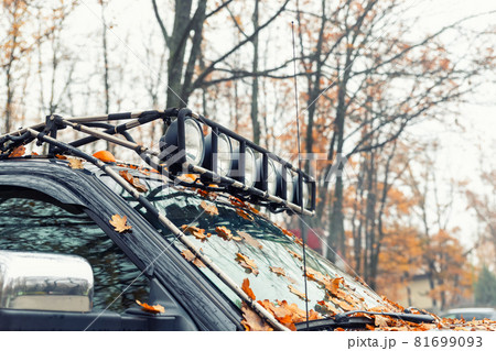 Close-up detail view of custom made roof rack bar with extra headlight mounted on roof of heavy duty pick up suv car against foggy autumn forest. Fallen leaves vehicle windshield. Fall weather drive Close-up detail view of custom made roof rack bar with extra headlight mounted on roof of heavy duty pick up suv car against foggy autumn forest. Fallen leaves vehicle windshield. Fall weather drive 81699093
