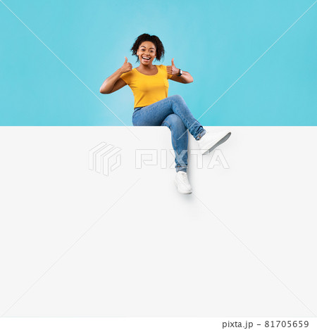 Excited young black lady sitting on blank white paper banner with mockup for ad, showing thumb up on blue background 81705659