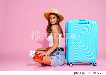 Smiling girl with a tickets money and passport are going to travel. Sitting near suitcase in shorts white top and straw hat. Pink background. looks at the camera 81709777