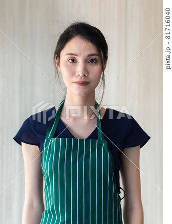Portrait Close up studio mug shot of young beautiful Asian professional female barista wears green stripe apron with black shirt stands look at camera in front brown wooden wallpaper background 81716040