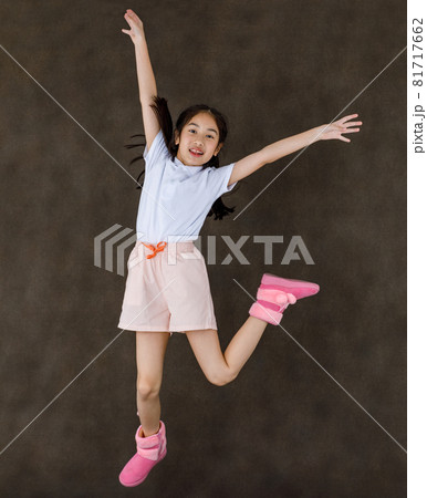 Portrait studio shot of little cute playful happy Asian girl wears white shirt and pink shorts and boots jumping high showing peace sign with two fingers overhead in front of brown wall background 81717662