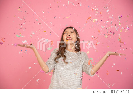 Portrait of a cheerful beautiful Asian womanl wearing dress standing standing under confetti rain and celebrating isolated over pink background 81718571