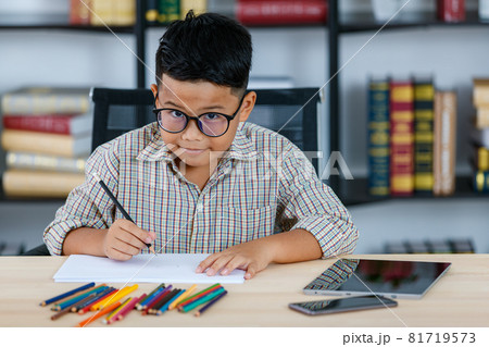 Lovely Asian boy wearing glasses and grey checked shirt sitting on desk of reading room and happily smiling while enjoy color drawing and art sketching on paper by blue pencil 81719573