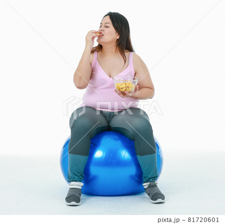 Isolated studio shot of Asian happy unhealthy overweight oversize girl in sport clothing leggings sit smiling on blue exercise fitball hold bowl of crunchy snack eating fast food on white background 81720601