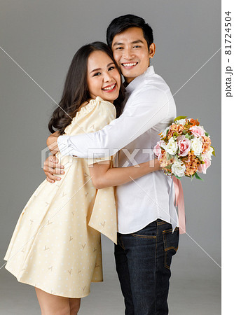 Portrait of young attractive Asian couple, man wearing white shirt, woman wearing beige dress hugging each other, woman holding bouquet of flower. Concept for pre wedding photography 81724504