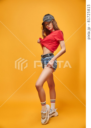 full body portrait sensual brunette wearing in jeans shorts red t-shirt and black cap. Looking at the camera in glasses. Yellow studio background. Girl in sneakers full body portrait sensual brunette wearing in jeans shorts red t-shirt and black cap. Looking at the camera in glasses. Yellow studio background. Girl in sneakers 81725815