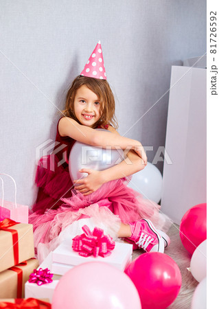 Little girl in pink dress and birthday hat sitting at the floor with many present gift box, balloons 81726592